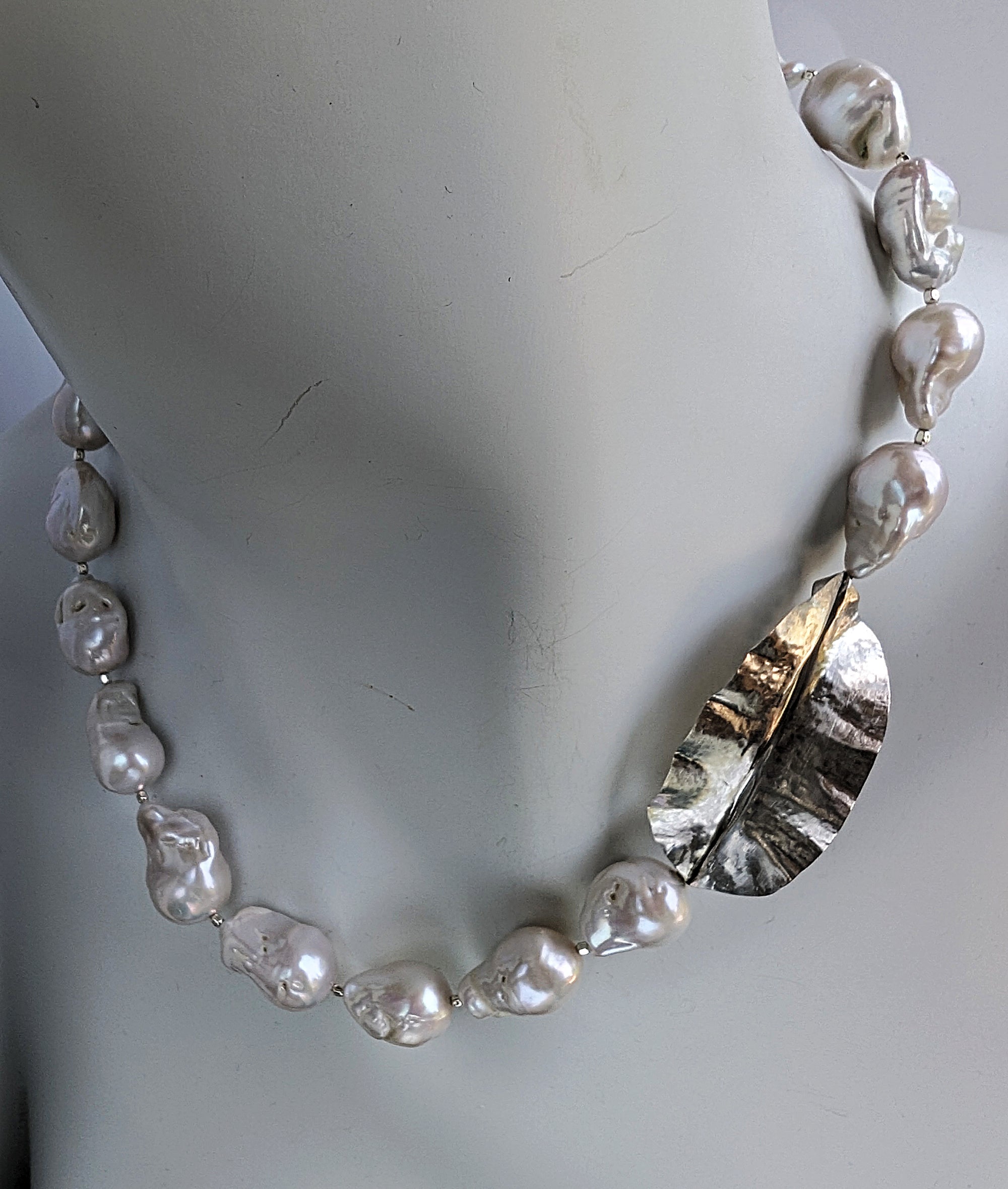 Featured on a white mannequin neck is a White Baroque pearl and silver necklace with hammered and fold form leaf design in silver with small silver beads in between each pearl.