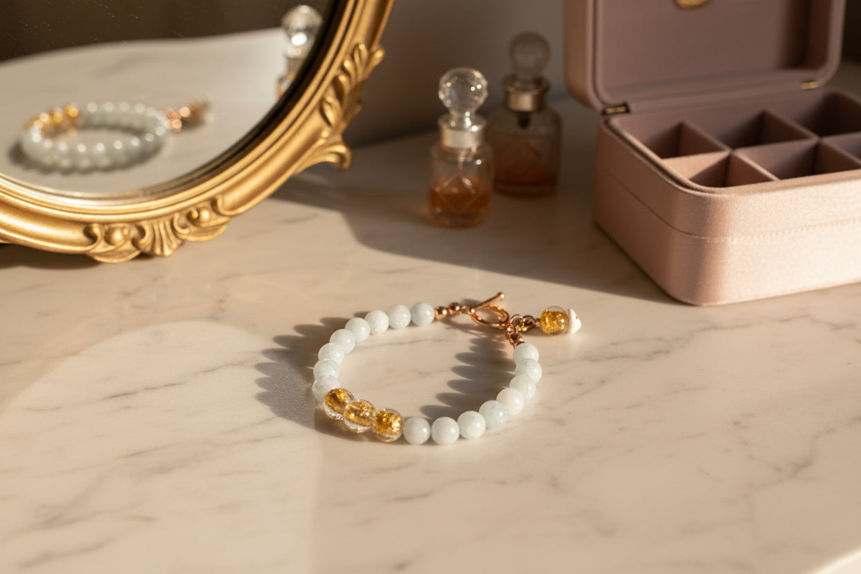 This picture tells a story of a light blue aquamarine and gold beaded bracelet on an elegant dressing table with gold mirror and reflected bracelet, two perfume bottles and the side of a jewelry chest.