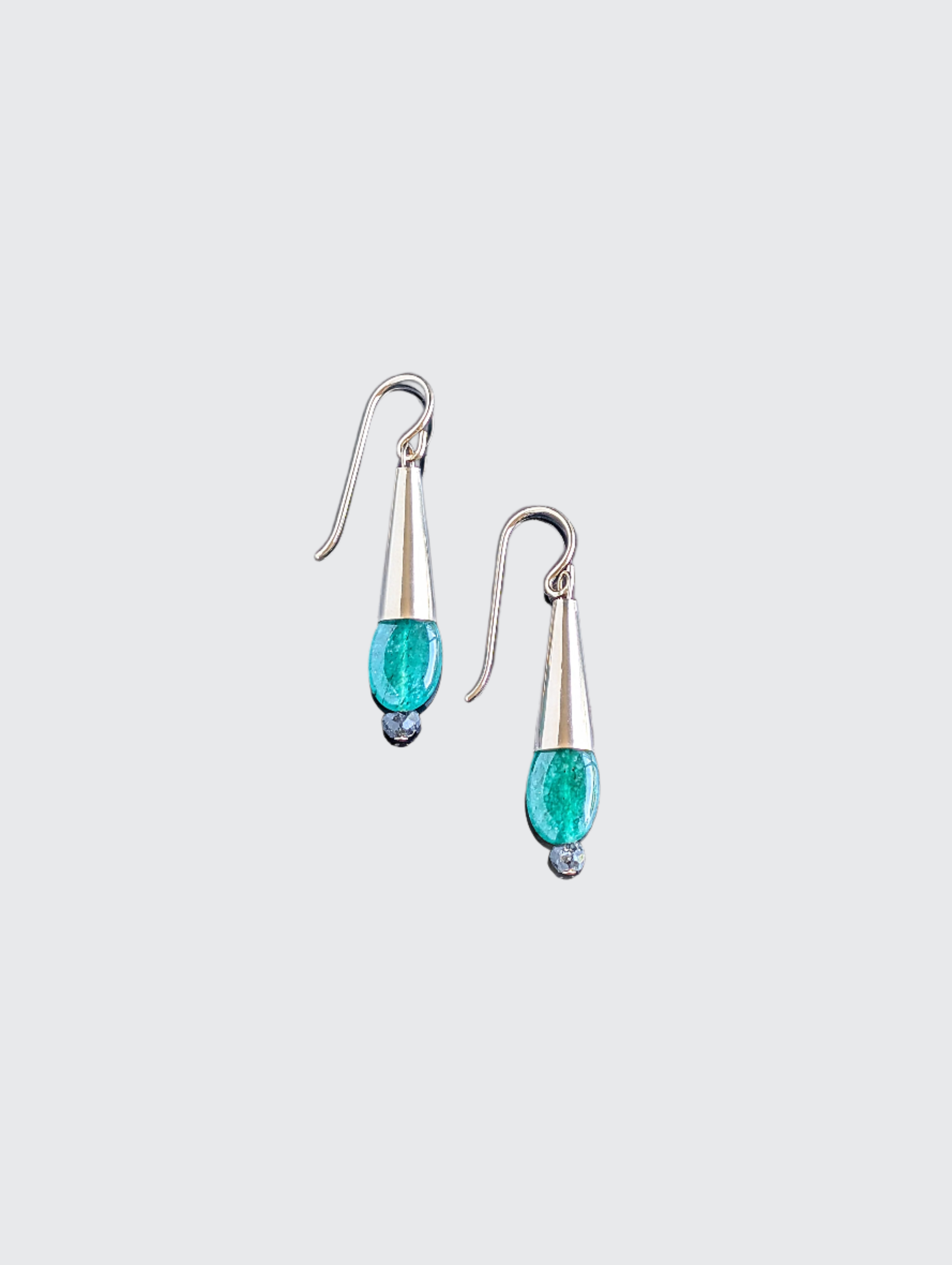 This image displays a pair of elegant Emerald Drop Earrings. The earrings feature a sleek, hanging design with each consisting of a polished metal droplet, from which a vibrant green emerald gemstone dangles like a delicate pendant.