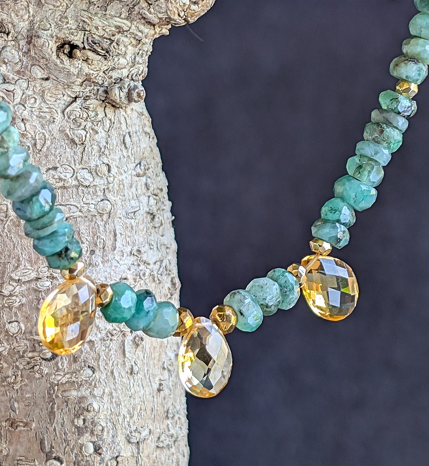A necklace with green emerald beads and faceted citrine briolette gems, displayed against a tree trunk.