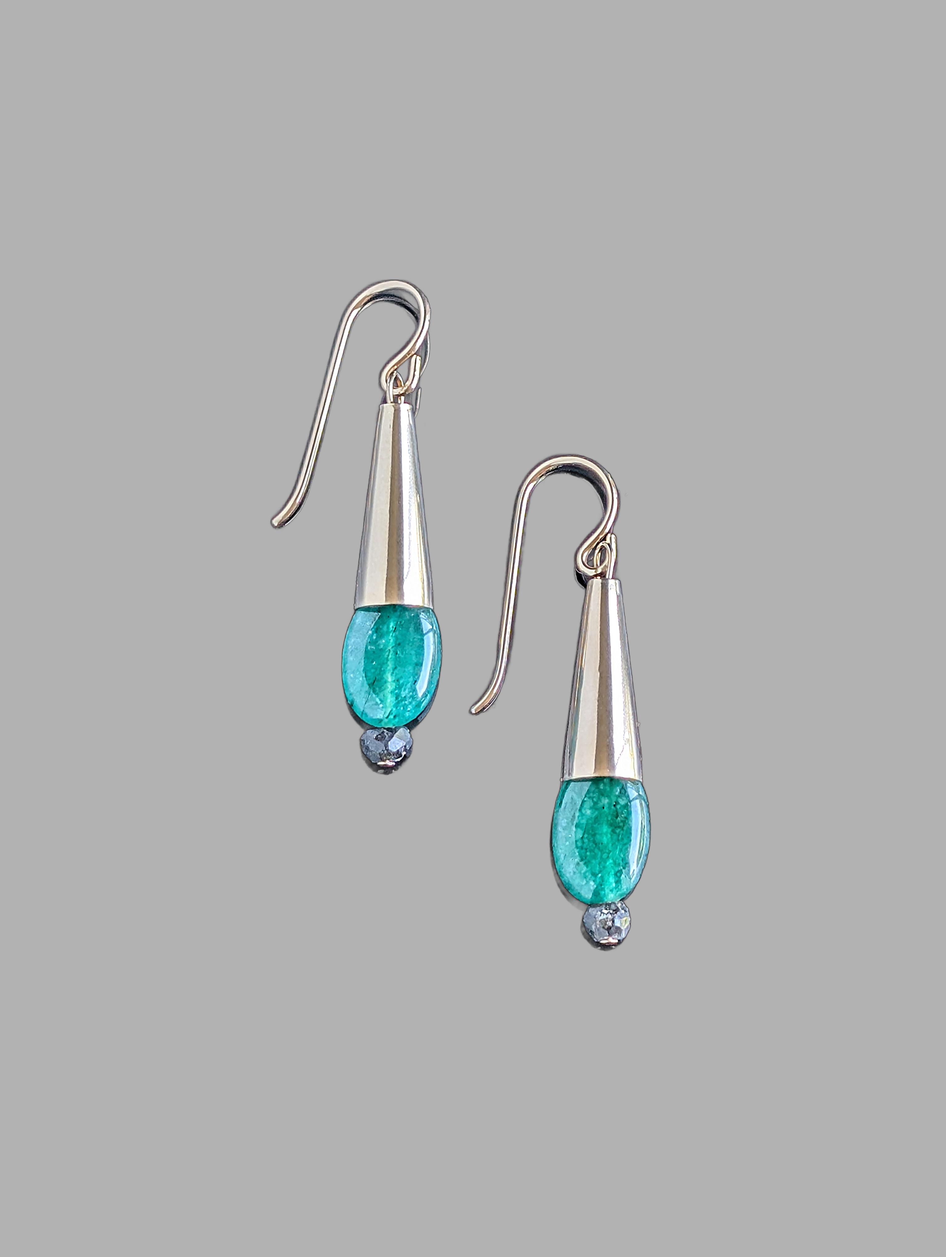 This image displays a pair of elegant Emerald Drop Earrings. The earrings feature a sleek, hanging design with each consisting of a polished metal droplet, from which a vibrant green emerald gemstone dangles like a delicate pendant.