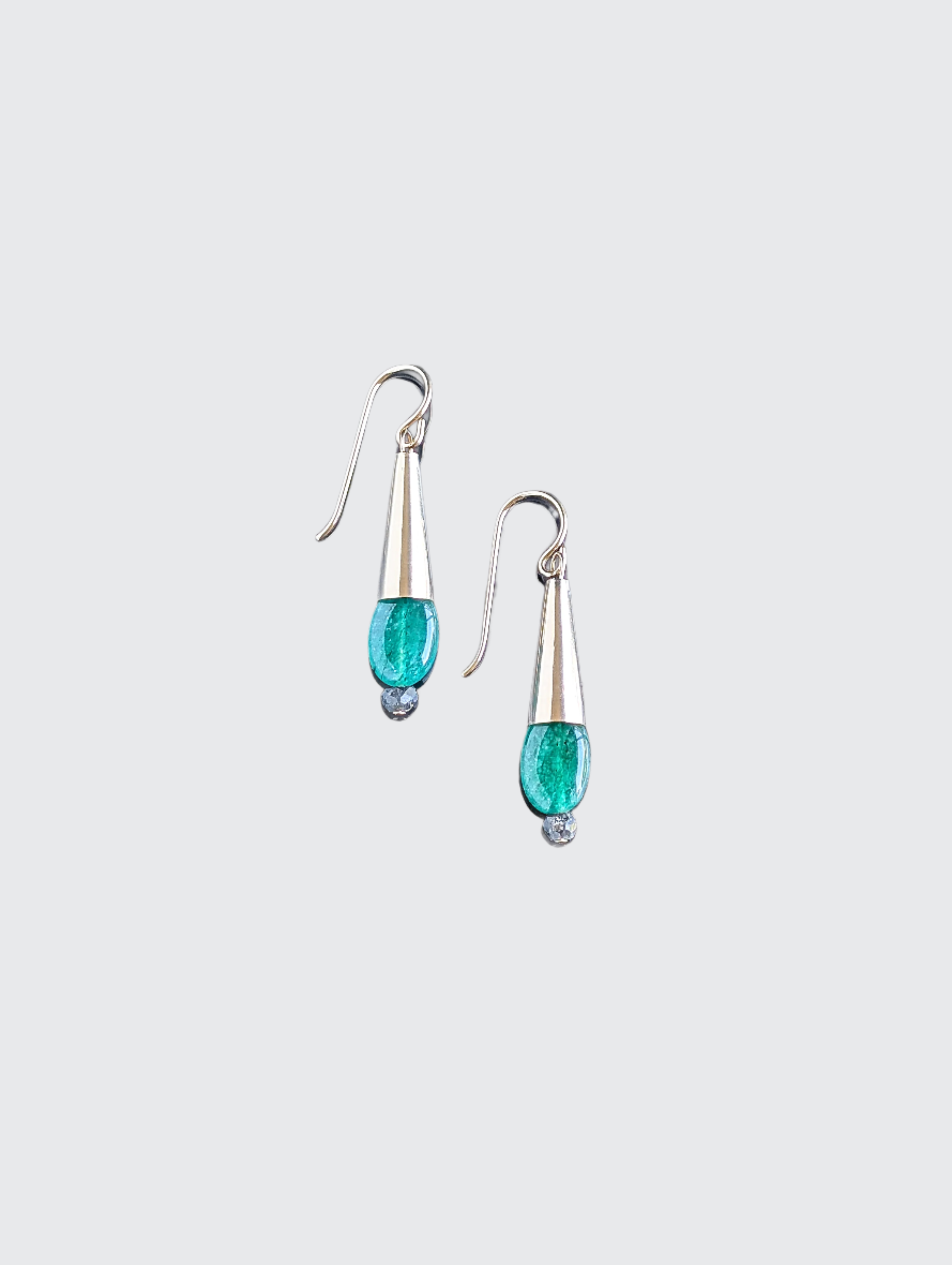 This image displays a pair of elegant Emerald Drop Earrings. The earrings feature a sleek, hanging design with each consisting of a polished metal droplet, from which a vibrant green emerald gemstone dangles like a delicate pendant.