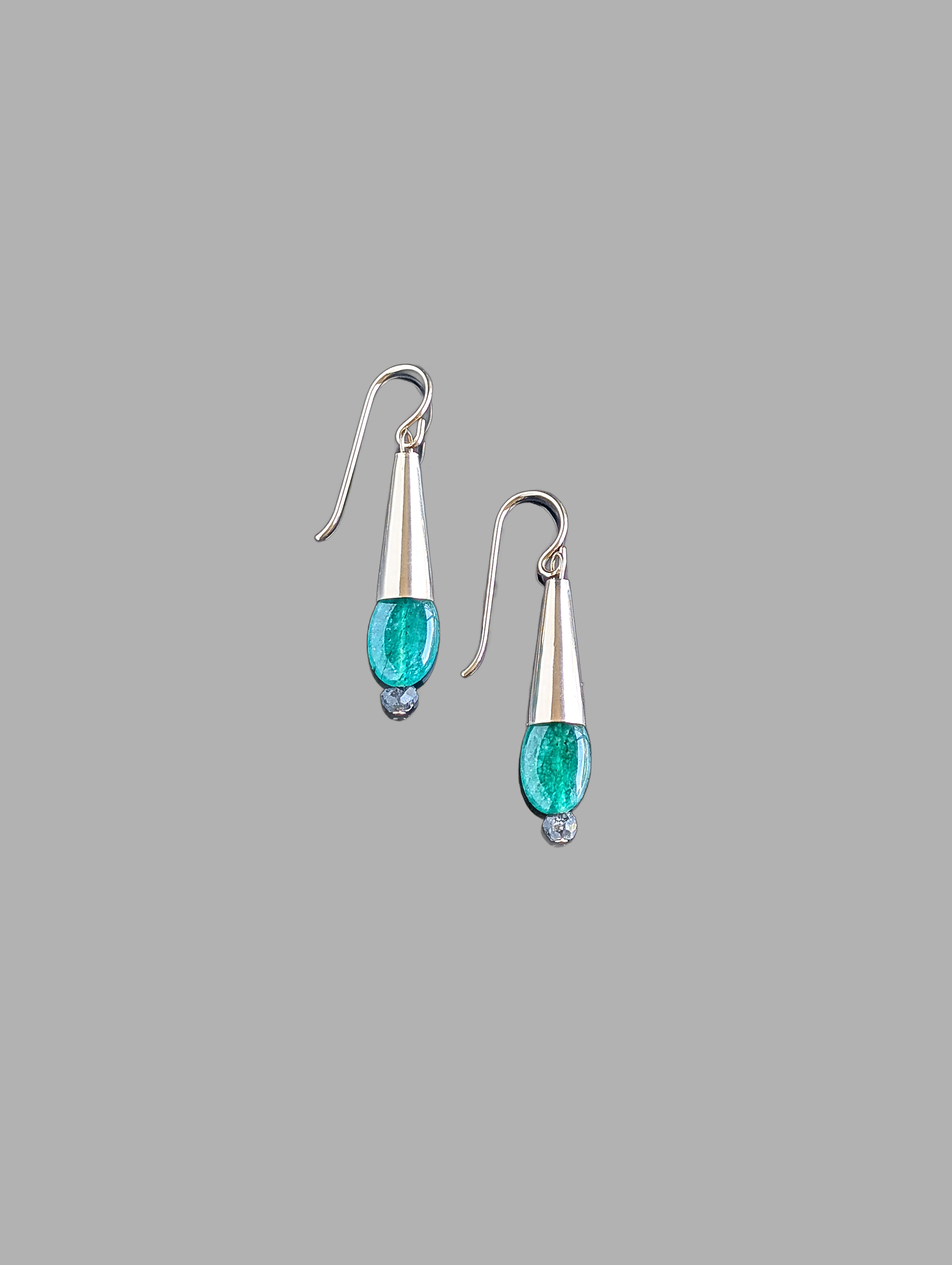 This image displays a pair of elegant Emerald Drop Earrings. The earrings feature a sleek, hanging design with each consisting of a polished metal droplet, from which a vibrant green emerald gemstone dangles like a delicate pendant.