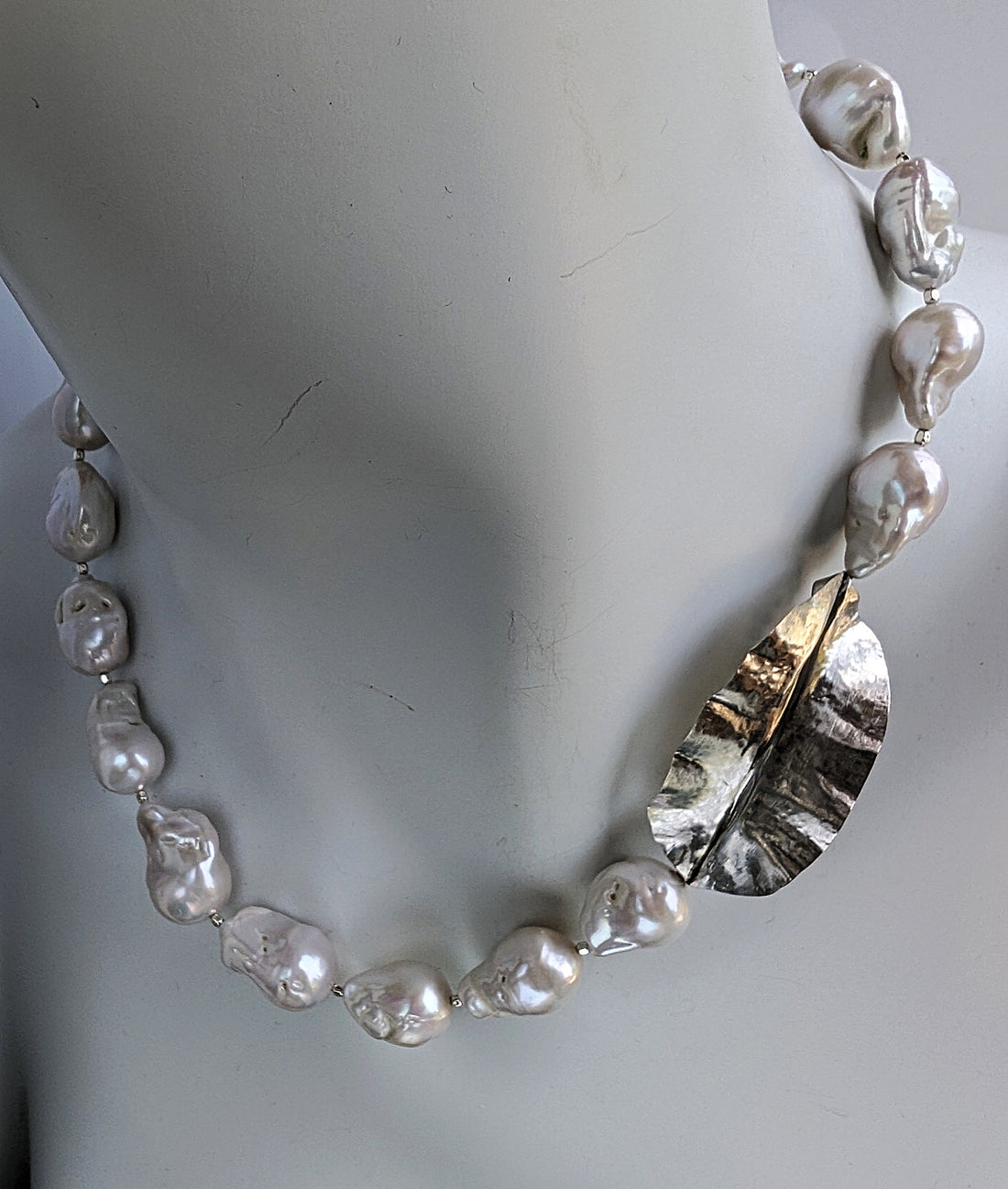 Featured on a white mannequin neck is a White Baroque pearl and silver necklace with hammered and fold form leaf design in silver with small silver beads in between each pearl.