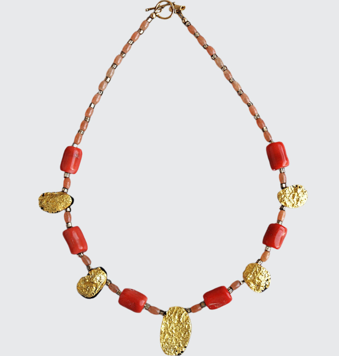 Showing the whole Gold and coral necklace with gold toggle clasp. There are five oval, tearshaped gold gilded textured tektite stones. The center stone is larger than the others. Inbetween are six dark orange barrel shaped and oval shaped coral beads with small gold glass beads inbetween and tapering to the clasp measuring 18".