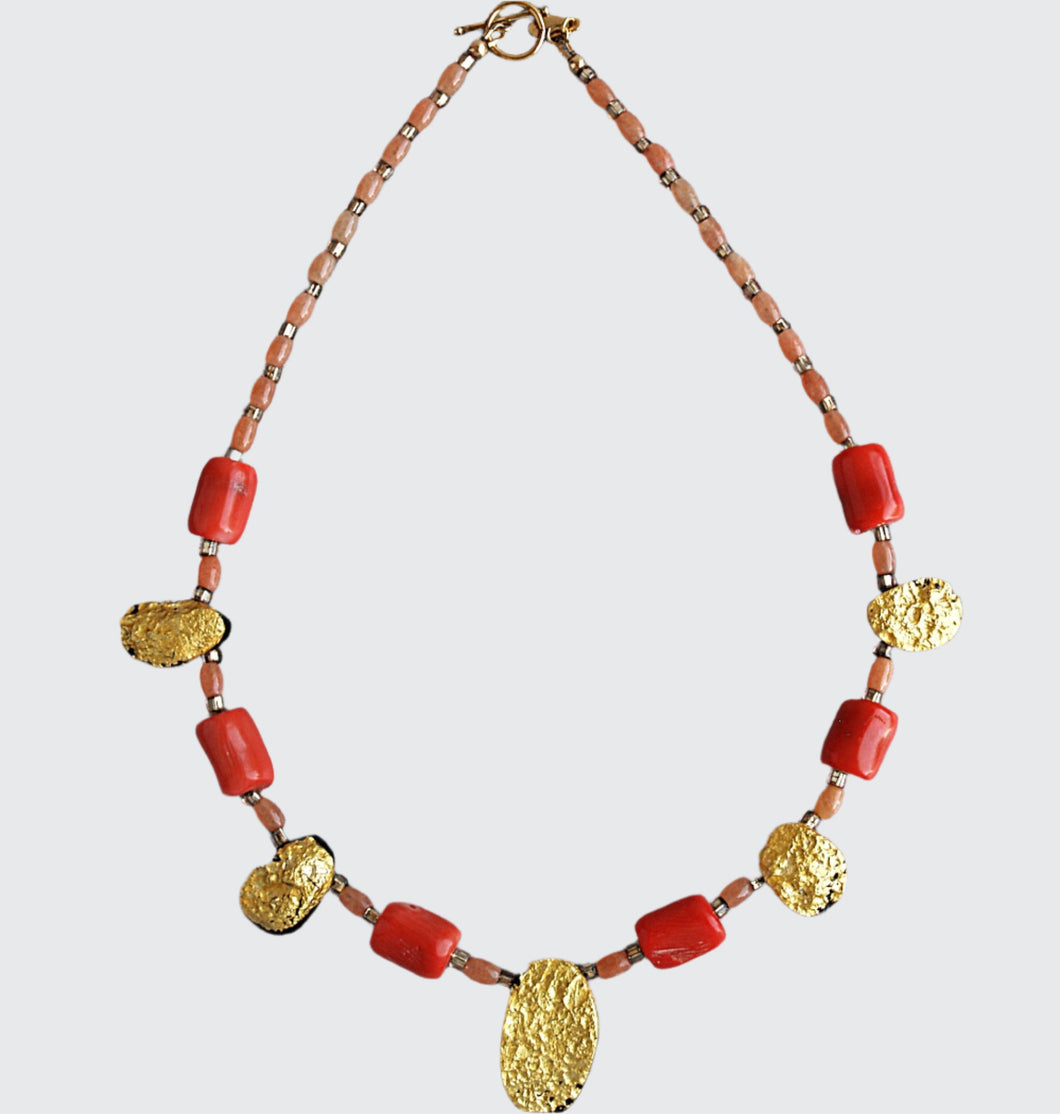 Showing the whole Gold and coral necklace with gold toggle clasp. There are five oval, tearshaped gold gilded textured tektite stones. The center stone is larger than the others. Inbetween are six dark orange barrel shaped and oval shaped coral beads with small gold glass beads inbetween and tapering to the clasp measuring 18