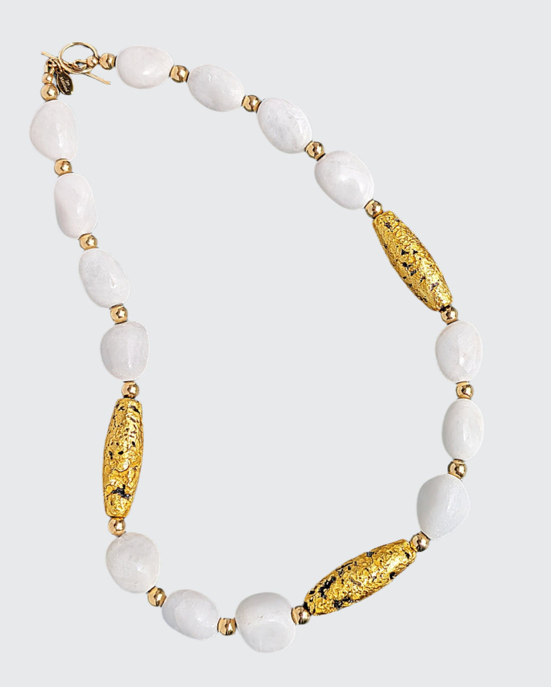 Enhance your allure with the &quot;Elegance In White&quot; Necklace featuring 23-Karat Gold Leaf on Lava Stone, White Aventurine, and Gold Beads.  Indulge in the enchantment of white and gold on your much-anticipated getaway. Each piece is meticulously crafted with hand-gilded, luminous 23-karat gold leaf on textured lava stone, complemented by radiant white aventurine and accented with shiny 14-Karat gold-filled toggle clasp and a distinctive designer signature tag.  Length: 19&quot; (48.26cm)