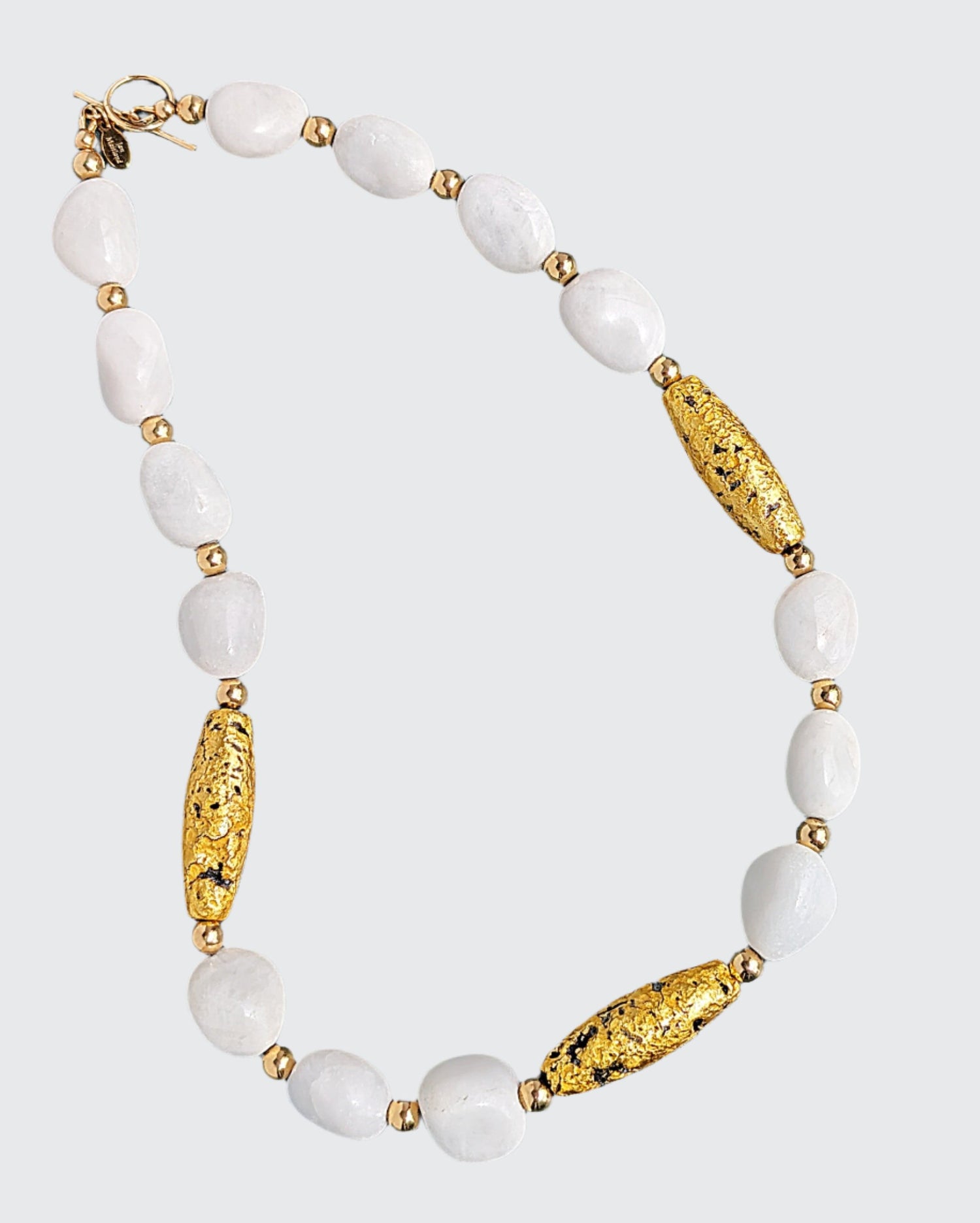 Enhance your allure with the &quot;Elegance In White&quot; Necklace featuring 23-Karat Gold Leaf on Lava Stone, White Aventurine, and Gold Beads.  Indulge in the enchantment of white and gold on your much-anticipated getaway. Each piece is meticulously crafted with hand-gilded, luminous 23-karat gold leaf on textured lava stone, complemented by radiant white aventurine and accented with shiny 14-Karat gold-filled toggle clasp and a distinctive designer signature tag.  Length: 19&quot; (48.26cm)