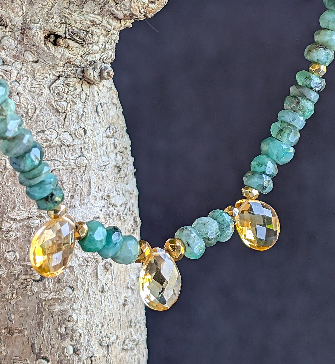 A necklace with green emerald beads and faceted citrine briolette gems, displayed against a tree trunk.