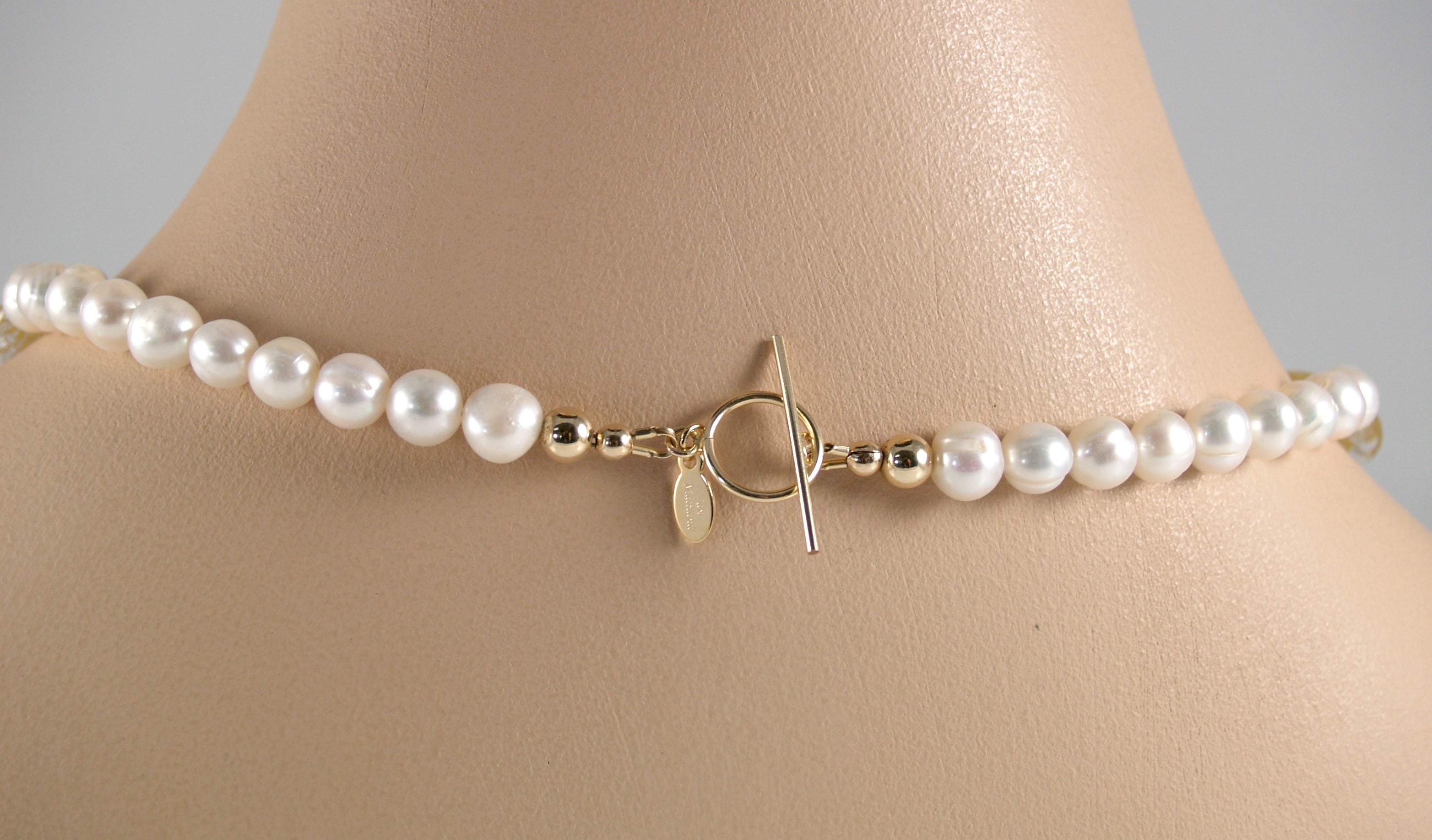 The back of white pearl necklace showing the gold toggle clasp and designer tag, featured on a mannequin neck. 